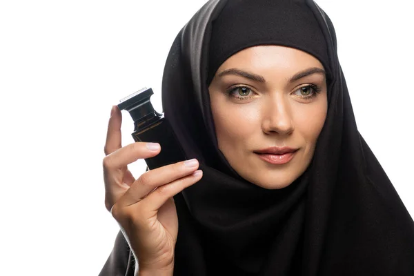 Joven musulmana en hiyab sosteniendo una botella de perfume y mirando hacia otro lado aislada en blanco - foto de stock