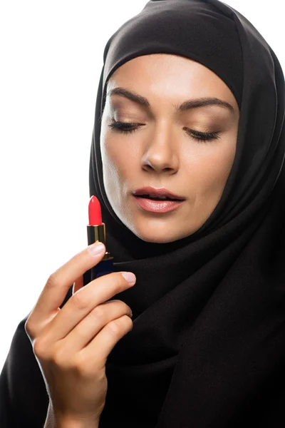 Mujer musulmana joven en hijab mirando lápiz labial rojo aislado en blanco - foto de stock