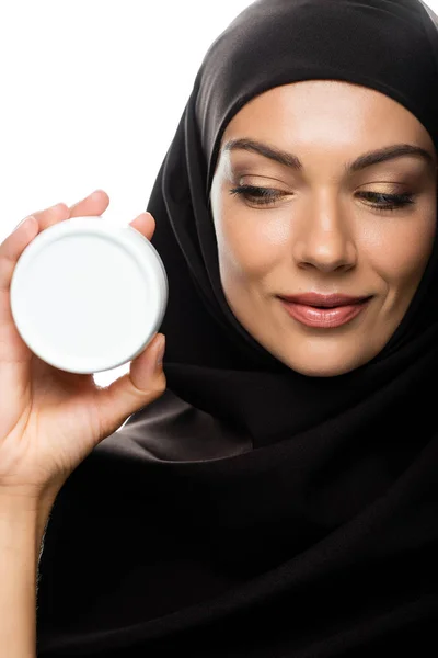 Smiling young Muslim woman in hijab holding container with cosmetic cream isolated on white — Stock Photo