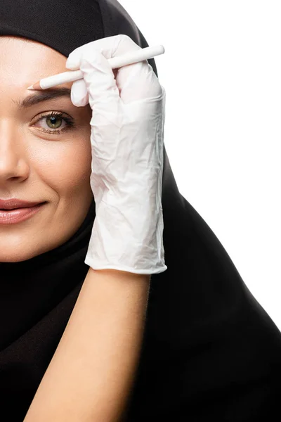 Vista recortada de cirujano plástico que marca la cara de mujer musulmana joven para cirugía plástica aislado en blanco - foto de stock