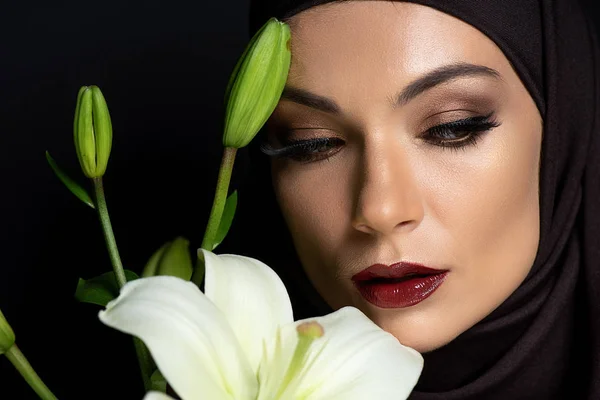 Schöne muslimische Frau im Hijab mit rauchigen Augen und roten Lippen, die Lilie isoliert auf schwarz hält — Stockfoto