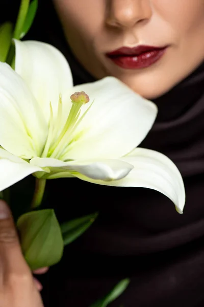 Cropped view of beautiful Muslim woman in hijab with red lips holding lily isolated on black — Stock Photo