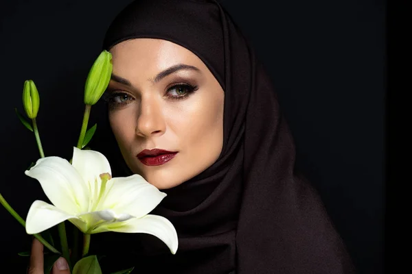 Bella donna musulmana in hijab con occhi fumosi e labbra rosse tenendo giglio isolato su nero — Foto stock