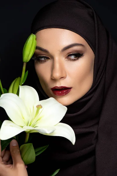 Красивая мусульманка в хиджабе с дымчатыми глазами и красными губами, держащая лилию изолированной на черном — стоковое фото