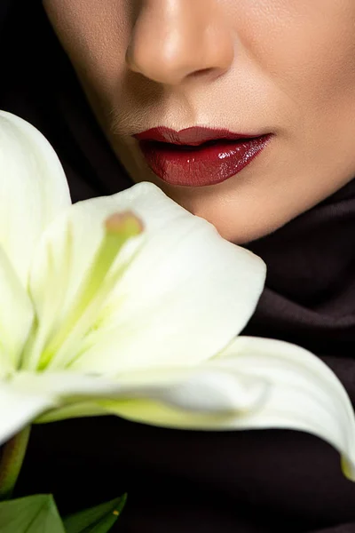 Vista parcial de la mujer musulmana en hiyab con labios rojos sosteniendo lirio aislado en negro - foto de stock