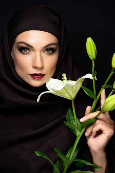 Attractive Muslim woman in hijab with red lips holding lily isolated on black — Stock Photo