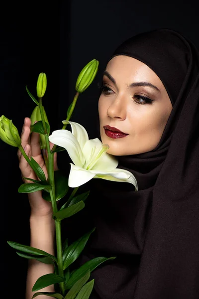 Attractive Muslim woman in hijab with red lips holding lily isolated on black — Stock Photo