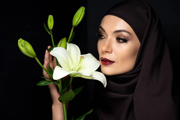 Attraente donna musulmana in hijab con labbra rosse tenendo giglio isolato su nero — Foto stock
