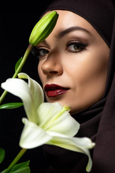 Portrait de femme musulmane séduisante en hijab avec lèvres rouges tenant le lis isolé sur noir — Photo de stock