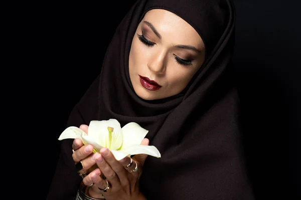 Beautiful Muslim woman in hijab in golden rings and bracelet holding lily isolated on black — Stock Photo