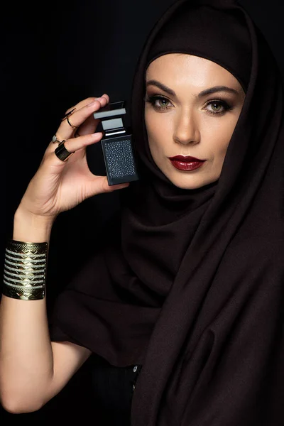 Hermosa mujer musulmana en hijab con maquillaje en joyas de oro celebración de perfume aislado en negro - foto de stock