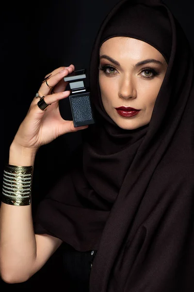 Beautiful Muslim woman in hijab with makeup in golden jewelry holding perfume isolated on black — Stock Photo