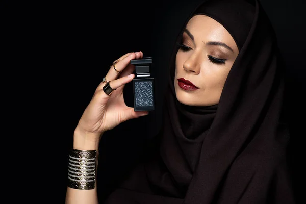 Beautiful Muslim woman in hijab with makeup in golden jewelry holding perfume isolated on black — Stock Photo