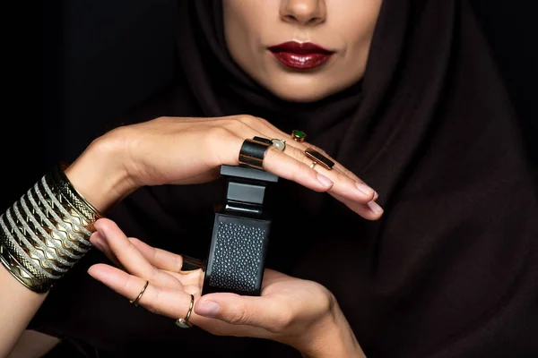 Vista recortada de la hermosa mujer musulmana en hijab con maquillaje en joyería de oro celebración de perfume aislado en negro - foto de stock