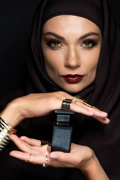 Hermosa mujer musulmana en hijab con maquillaje en joyas de oro celebración de perfume aislado en negro - foto de stock