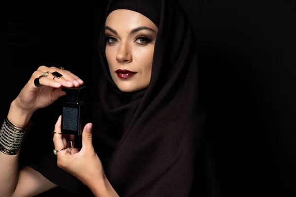 Bela mulher muçulmana em hijab com maquiagem em jóias douradas segurando perfume isolado em preto — Fotografia de Stock