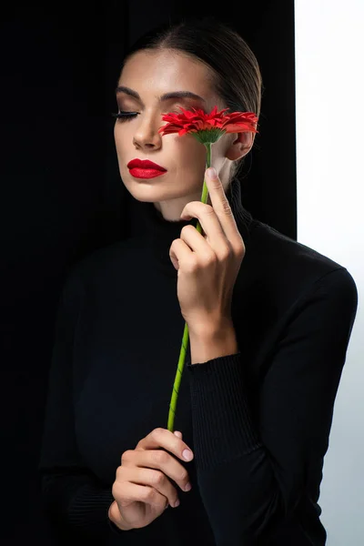 Hermosa mujer con labios rojos y ojos cerrados sosteniendo gerberas rojas aisladas en blanco y negro - foto de stock