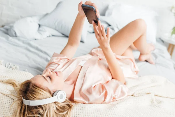 Selective focus of happy girl using smartphone and listening music while lying on bed — Stock Photo