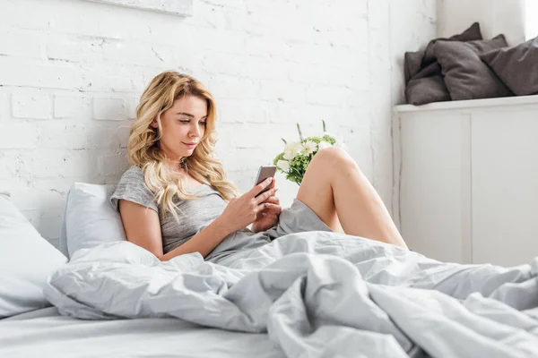 Attractive girl using smartphone near bedside table with flowers at home — Stock Photo