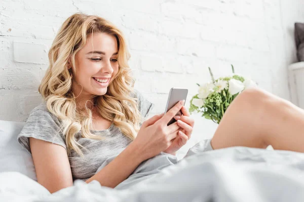 Selective focus of cheerful woman using smartphone in bed — Stock Photo