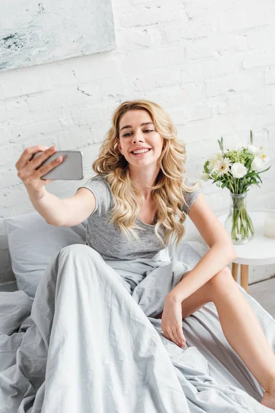 Selective focus of cheerful girl taking selfie near vase with flowers on bedside table — Stock Photo