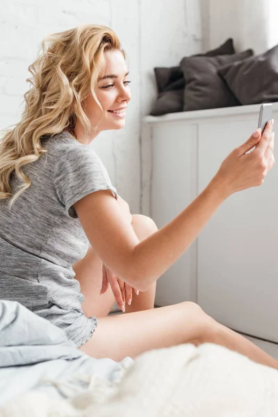 Selektiver Fokus einer glücklichen jungen Frau, die auf dem Bett sitzt und ihr Smartphone in der Hand hält — Stockfoto