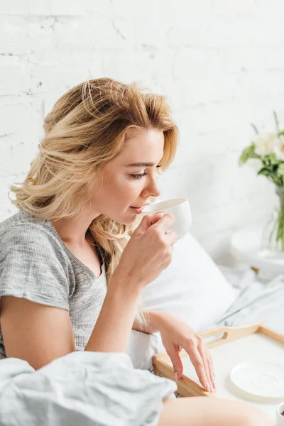 Selektiver Fokus des schönen blonden Mädchens, das eine Tasse Kaffee im Bett hält — Stockfoto