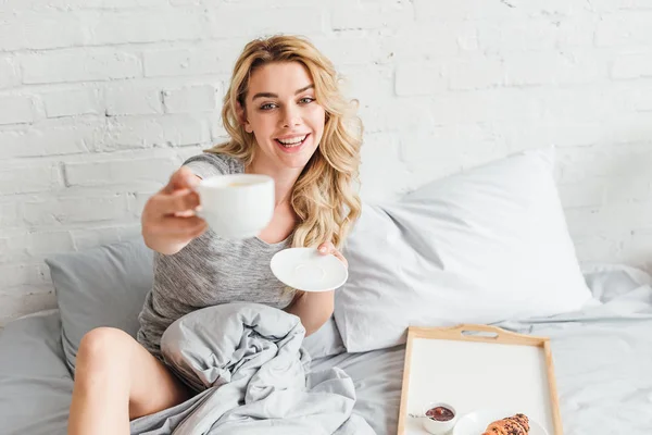 Избирательный фокус счастливой женщины, держащей чашку кофе и блюдце в постели — стоковое фото