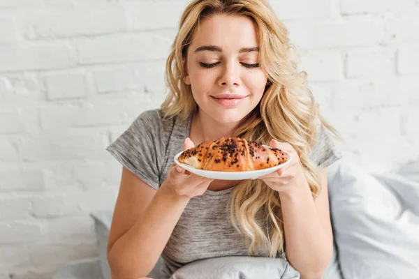 Donna felice guardando gustoso croissant sul piatto in camera da letto — Foto stock