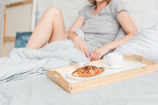 Foco seletivo de bandeja de madeira com croissant saboroso e café perto de mulher na cama — Fotografia de Stock