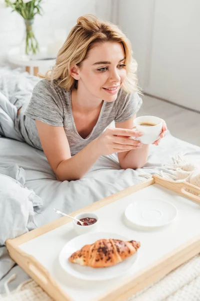 Fuoco selettivo di ragazza allegra che tiene la tazza di caffè vicino a vassoio con croissant saporito su letto — Foto stock