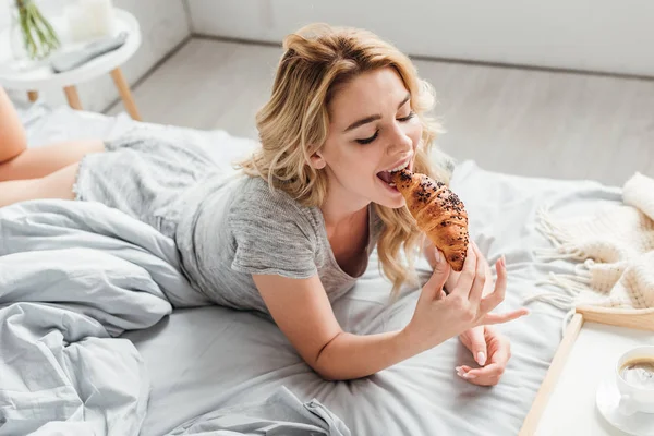 Над головой вид счастливой девушки, кушающей круассан возле подноса с чашкой кофе на кровати — стоковое фото