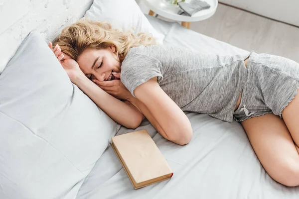 Overhead view of cheerful woman covering face with laughing and lying on bed near book — Stock Photo