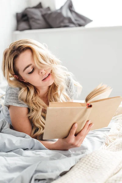 Enfoque selectivo de la mujer joven y atractiva lectura de libro mientras está acostado en la cama - foto de stock