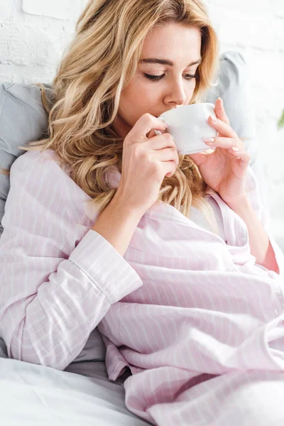 Attraktive Frau trinkt Kaffee, während sie Tasse im Bett hält — Stockfoto
