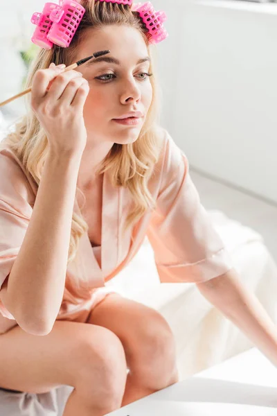 Pretty woman in pink hair curlers styling eyebrows in bedroom — Stock Photo