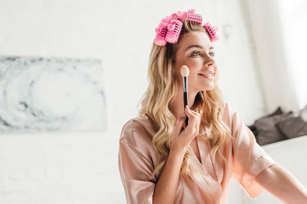 Mulher alegre em encrespadores de cabelo rosa segurando escova cosmética perto do rosto no quarto — Fotografia de Stock