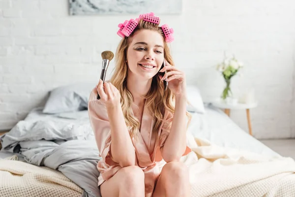 Cheerful girl holding cosmetic brush while talking on smartphone — Stock Photo