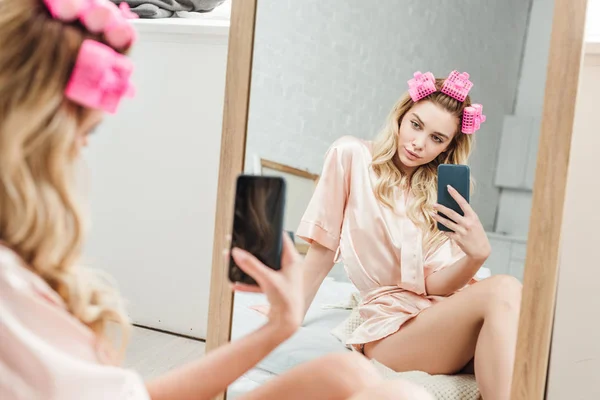 Foco seletivo da mulher atraente com encrespadores de cabelo segurando smartphone enquanto tira foto perto do espelho — Fotografia de Stock