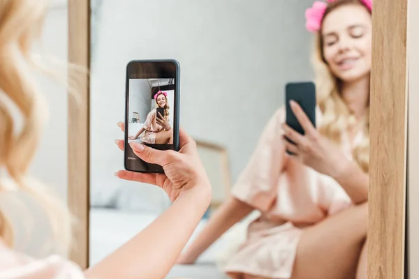 Вибірковий фокус усміхненої жінки з лижниками для волосся, що тримає смартфон, роблячи фотографії біля дзеркала — стокове фото