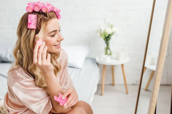 Selective focus of happy girl touching pink hair curlers and looking at mirror — Stock Photo