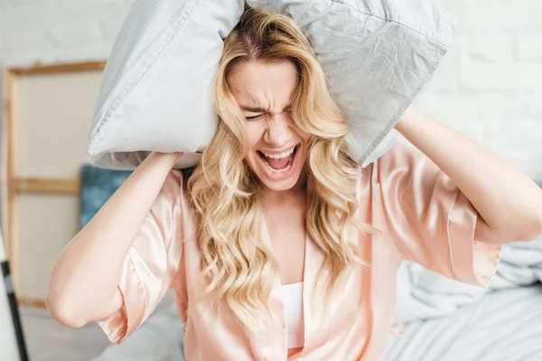 Emotional woman in silk robe holding pillow screaming in bedroom — Stock Photo