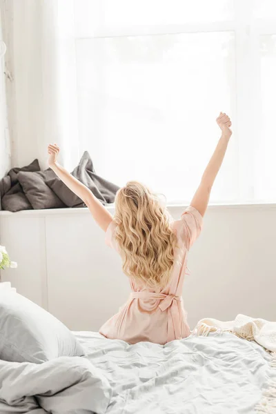 Вид сзади блондинки с вытянутыми руками, сидящей на кровати — стоковое фото