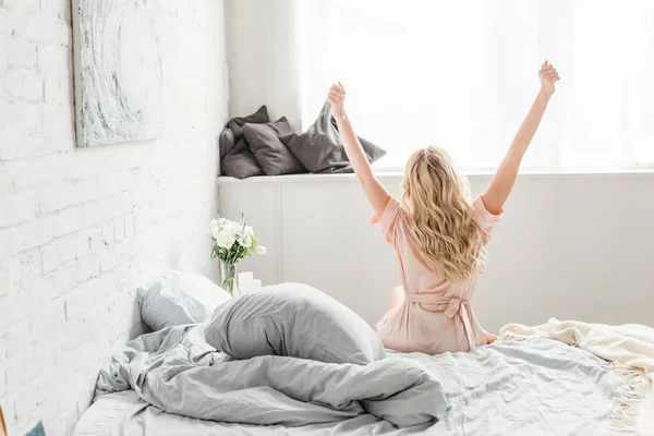 Rückansicht einer jungen Frau mit ausgestreckten Händen, die auf dem Bett sitzt — Stockfoto