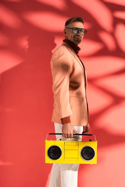 Homme heureux et élégant dans des lunettes de soleil tenant boombox jaune sur rouge avec des ombres — Photo de stock