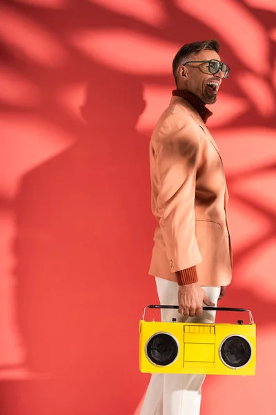 Cheerful and stylish man in sunglasses holding yellow boombox on red with shadows — Stock Photo