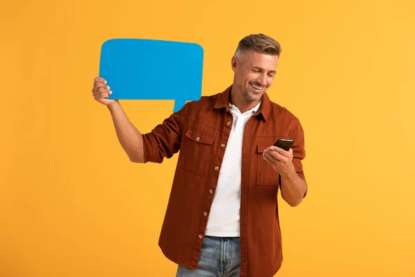 Hombre feliz sosteniendo burbuja de habla azul y teléfono inteligente aislado en naranja - foto de stock
