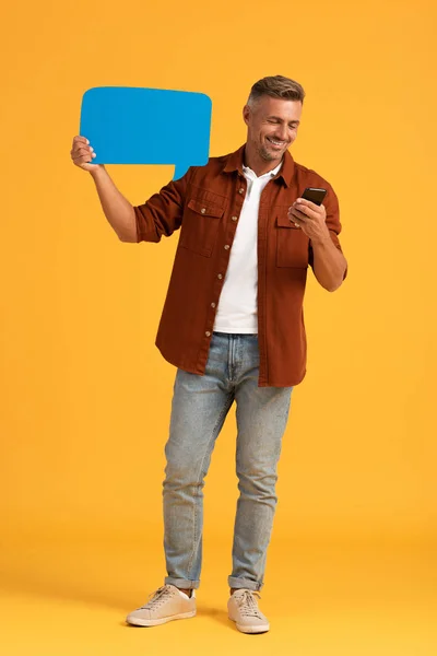 Hombre feliz sosteniendo burbuja de habla azul y teléfono inteligente en naranja - foto de stock