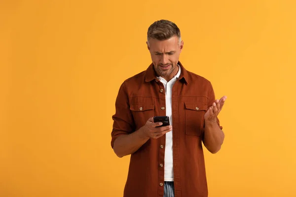 Недовольный человек смотрит на смартфон и жесты изолированы на оранжевый — стоковое фото