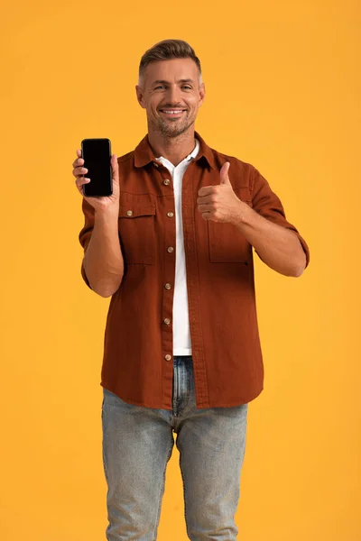Щасливий чоловік показує великий палець і тримає смартфон з порожнім екраном ізольовано на помаранчевому — стокове фото